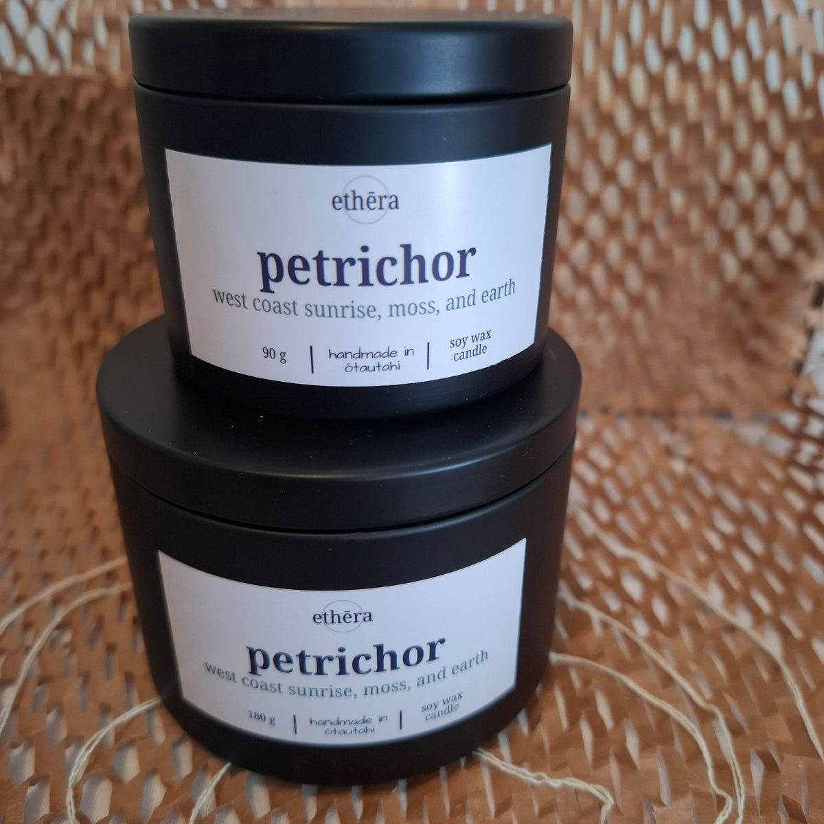 Two stacked black jars of ethéra Petrichor soy wax candles on a woven wicker surface. The lower jar is 180 g and the upper jar is 90 g, both with white labels featuring the brand name 'ethéra' and scent name 'petrichor'.
