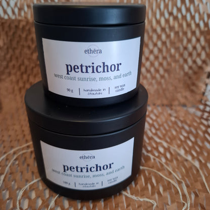 Two stacked black jars of ethéra Petrichor soy wax candles on a woven wicker surface. The lower jar is 180 g and the upper jar is 90 g, both with white labels featuring the brand name 'ethéra' and scent name 'petrichor'.