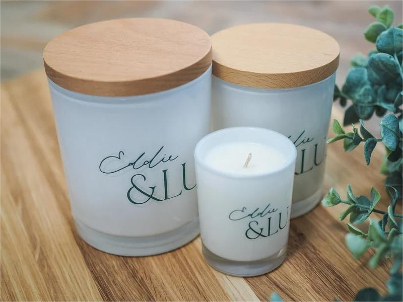 EL cocowax candle trio -  white jars, two largest lidded-wooden, on wood-grain table with fresh leaves.