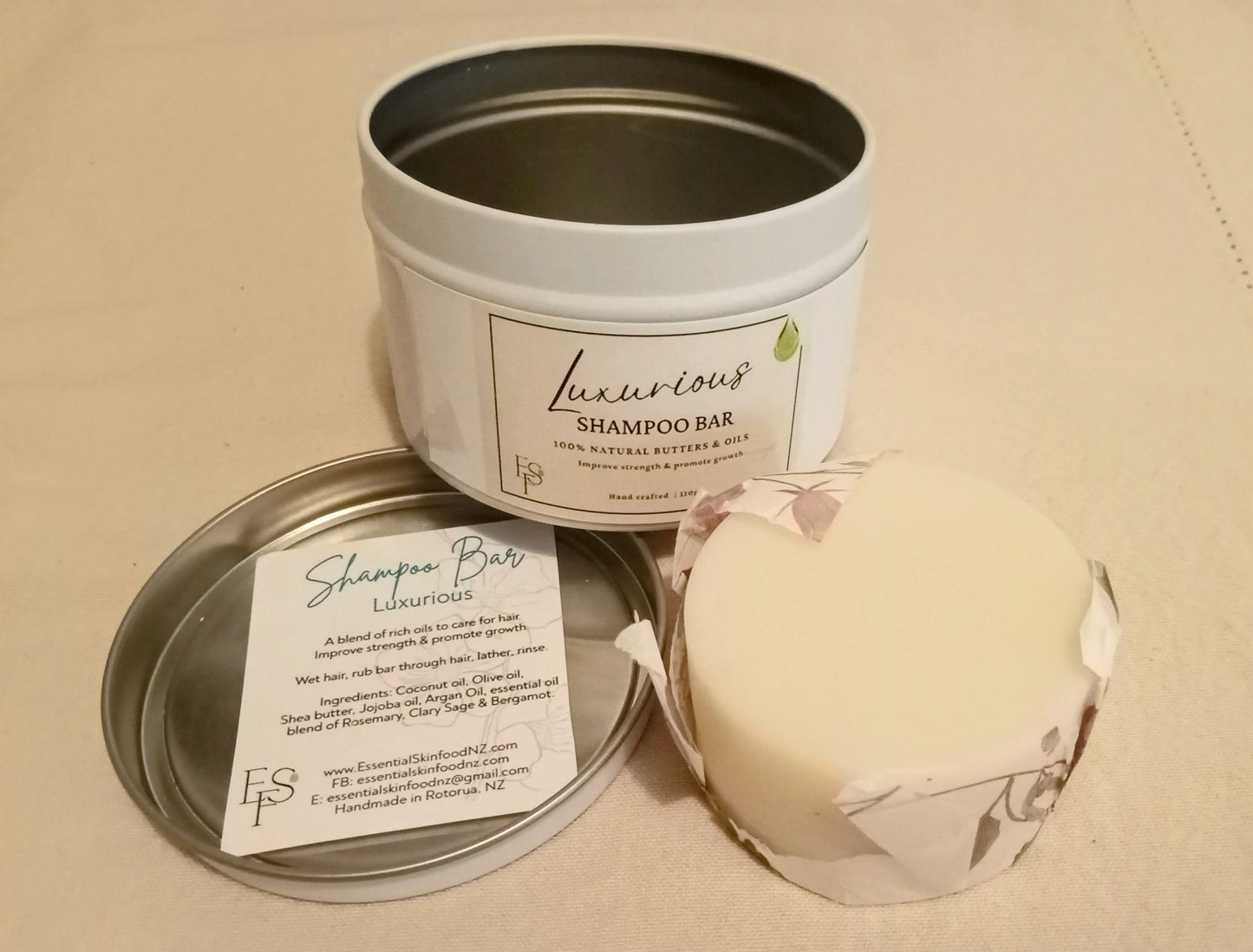 Image of small white round tin with paper label, a shampoo bar, and paper inset about product on beige linen background.