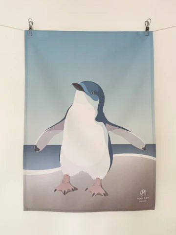 Tea-towel with blue penguin on a beach with a gradient sky print, clipped on line over white background