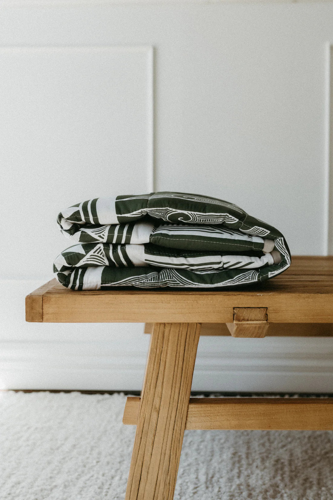 Folded blanket with a green and white pattern on a wooden bench against a white wall.
