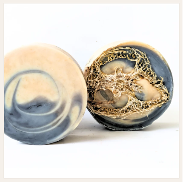 Two round blue & white soaps (same - front & back views) shown open on a white background.  Front view of  soap shows slice of loofah encased;.rear view shows detail of spiralled colour. mix.