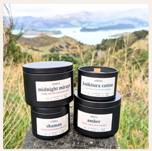 Four white soy wax candles in matte metal tins with white labels are arranged on a stone surface outdoors, with a scenic mountain and lake landscape in the background. The labels show the brand 'ethéra' and scent names including 'midnight mirror', 'kaikóra cotton', 'shaman', and 'amber'.