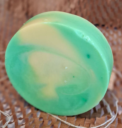 A round handmade soap bar with a marbled green and pale yellow swirl pattern, resting on a brown woven straw mat.