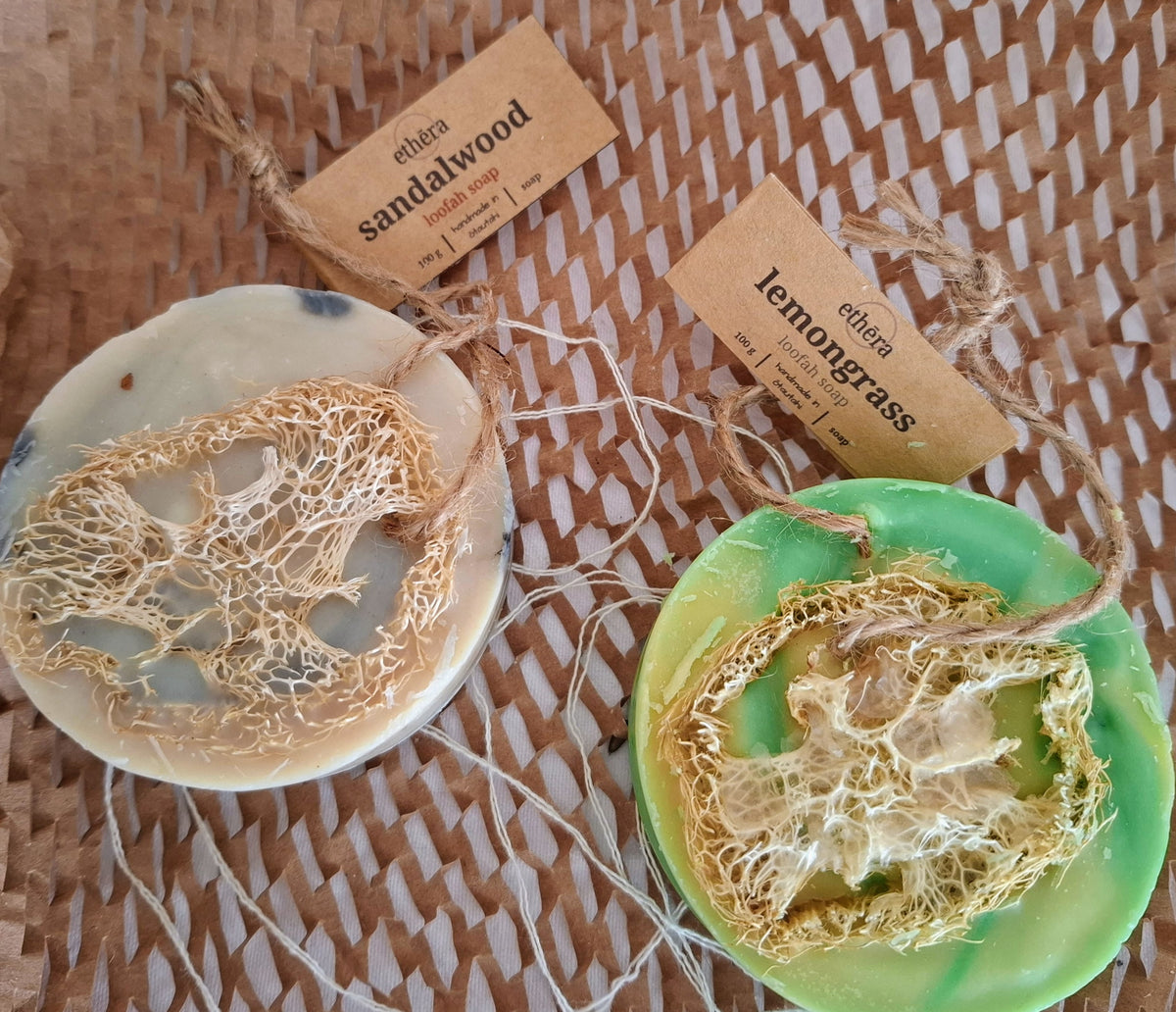 Two handmade soap bars with natural lather centers, one made from Sandalwood and the other from lemongrass, resting on a woven straw mat. Each soap is tied to a kraft paper tag with the brand name 'ethera.