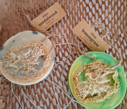Two handmade soap bars with natural lather centers, one made from Sandalwood and the other from lemongrass, resting on a woven straw mat. Each soap is tied to a kraft paper tag with the brand name 'ethera.