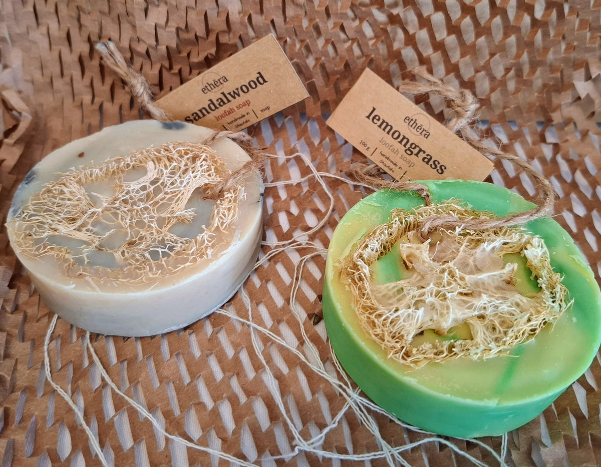 Two handmade loofah soap bars on a woven brown mat. One bar is light beige with a natural loofah overlay and a tag reading 'ebena sandalwood'.