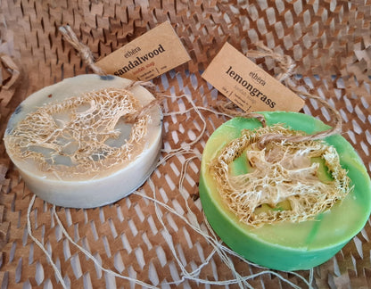 Two handmade loofah soap bars on a woven brown mat. One bar is light beige with a natural loofah overlay and a tag reading 'ebena sandalwood'.