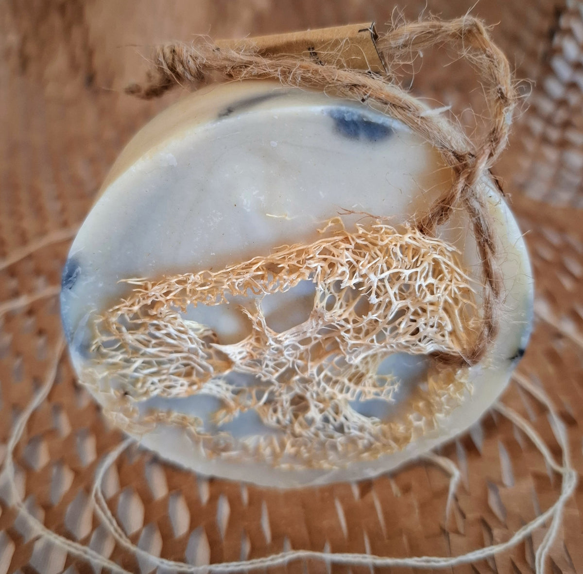 Blue & white round soap bar with loofah slice encased placed on natural weave & spiralled string.