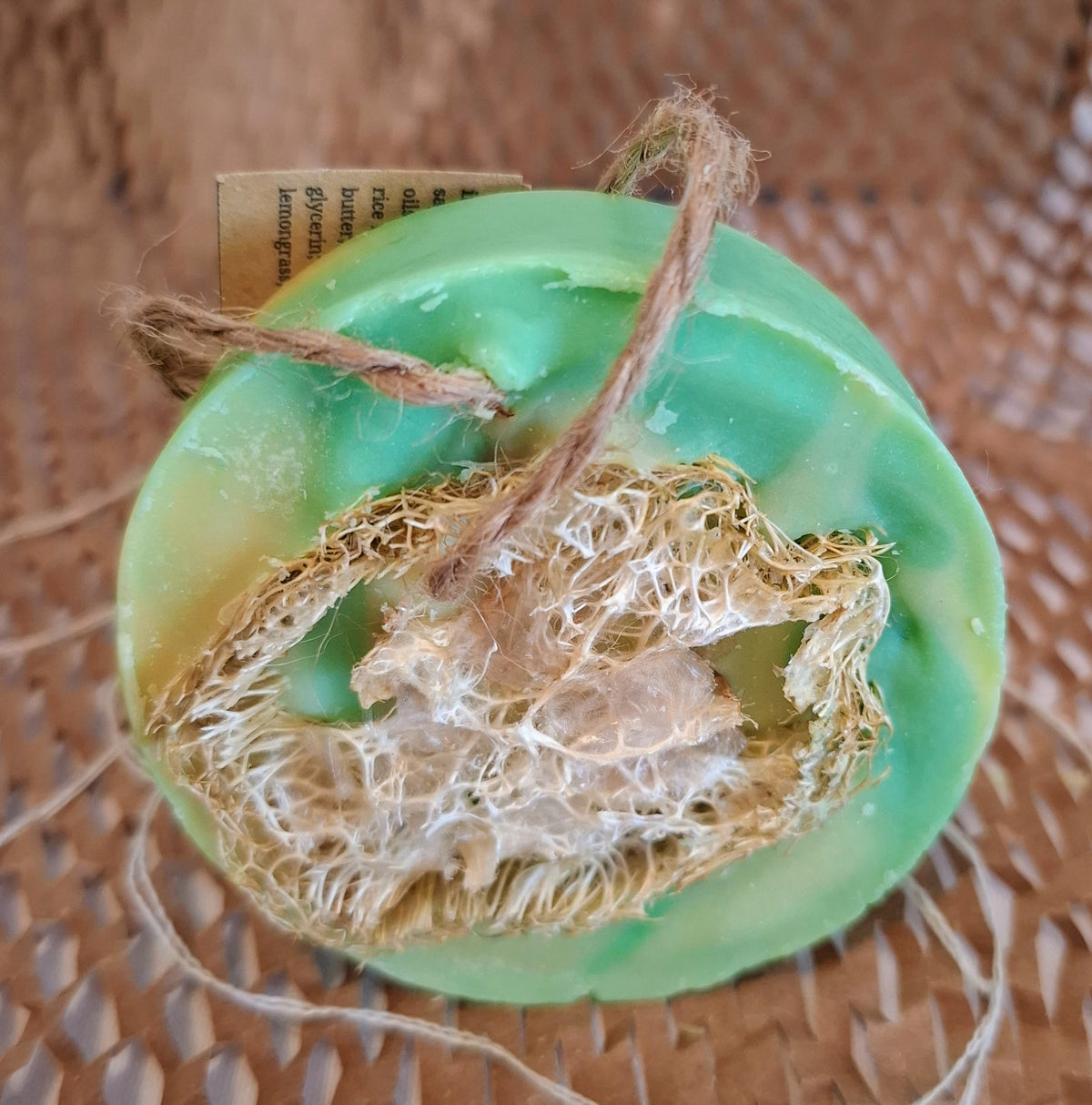 A green loofah soap bar with a natural loofah sponge attached and a small paper tag, shown on a brown woven mat background.