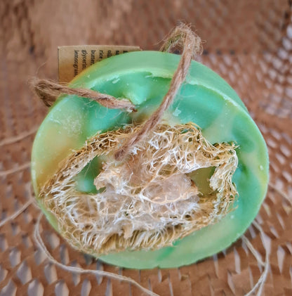 A green loofah soap bar with a natural loofah sponge attached and a small paper tag, shown on a brown woven mat background.