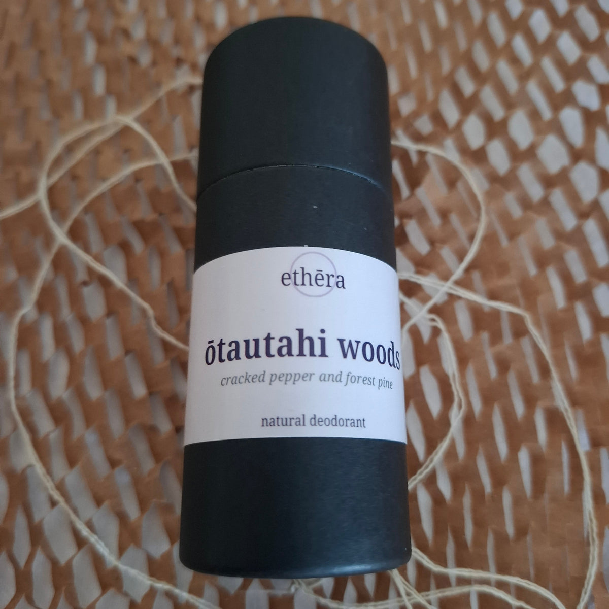 A black cylindrical deodorant stick with a white label reading 'ethéra ōtautahi woods cracked pepper and pine peat natural deodorant', placed on a woven brown surface with string.