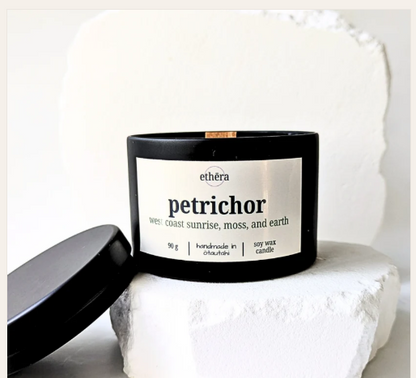 A white soy wax candle jar with a white label reading 'ethéra petrichor' sits on a white stone pedestal with its lid leaning beside it against a soft white background.