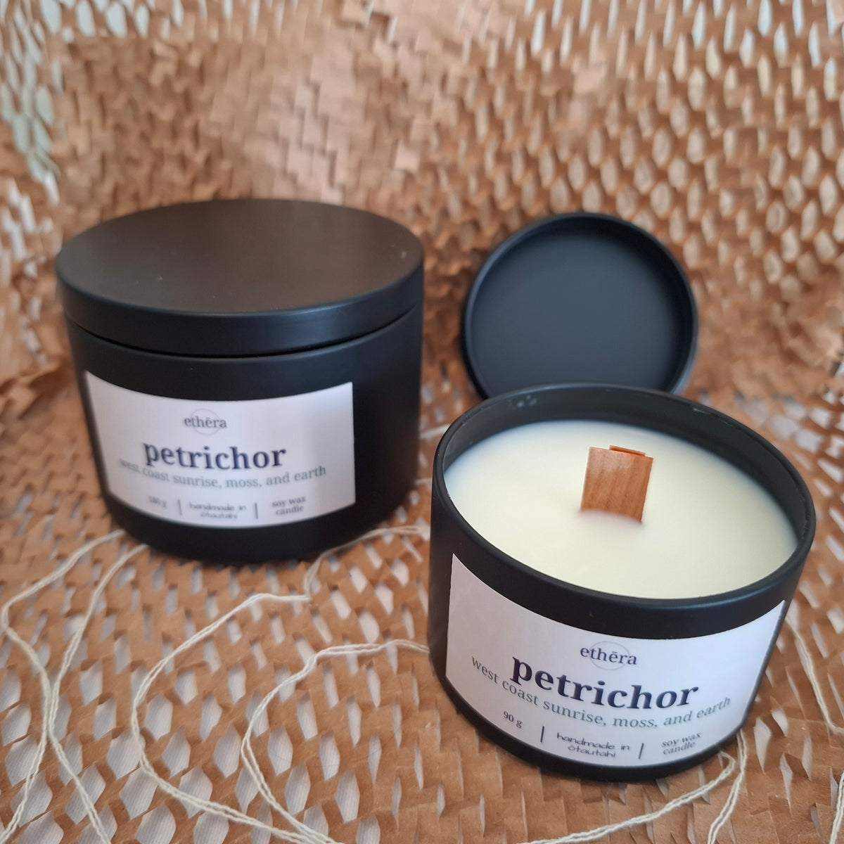 Two black metal candle tins on a woven straw surface, one closed and one open showing white wax with a wooden wick. The labels read ethera and petrichor.