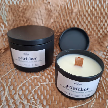 Two black metal candle tins on a woven straw surface, one closed and one open showing white wax with a wooden wick. The labels read ethera and petrichor.